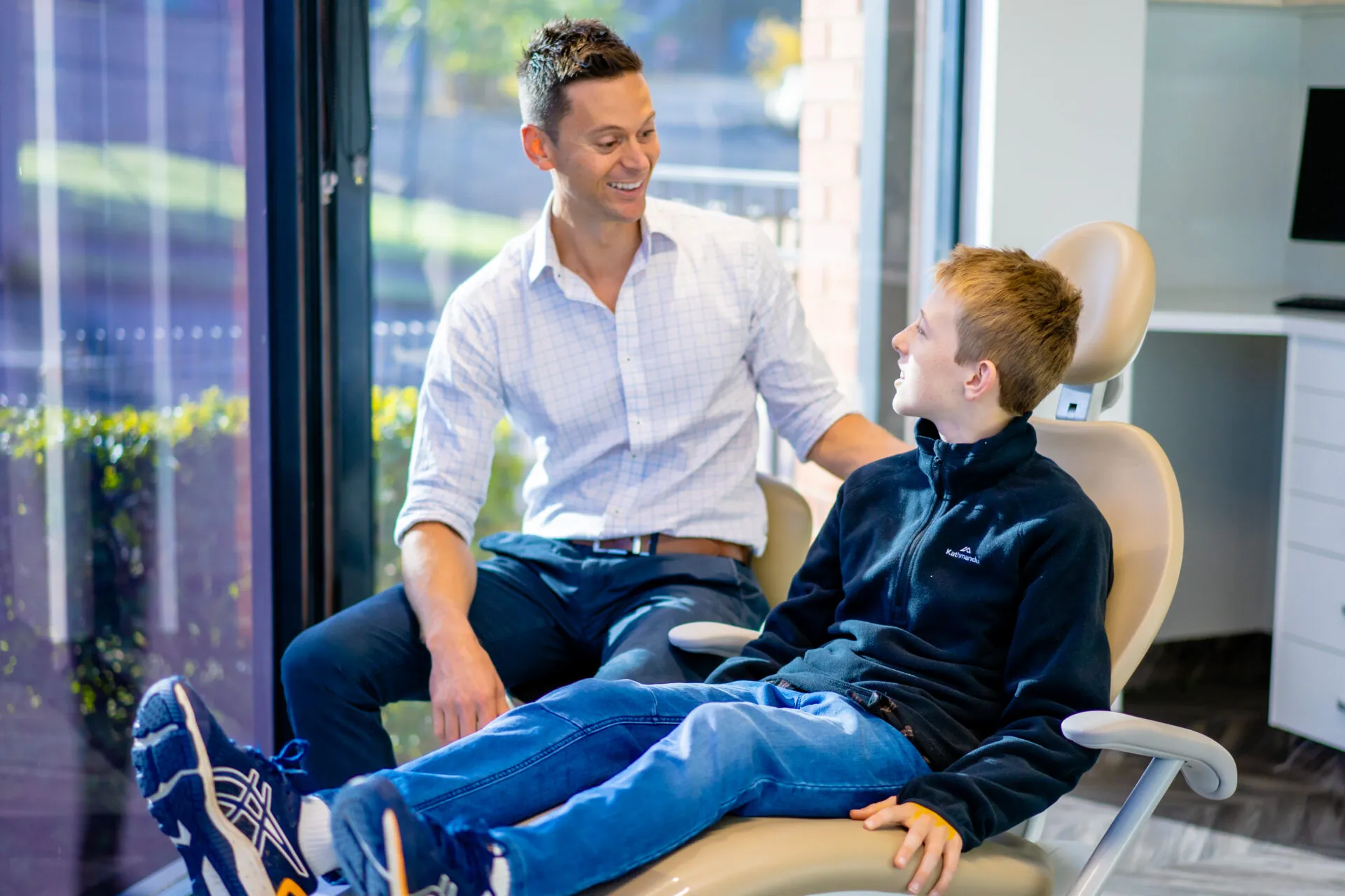 Orthodontist talking with a teenage boy seated in the treatment chair beside the window