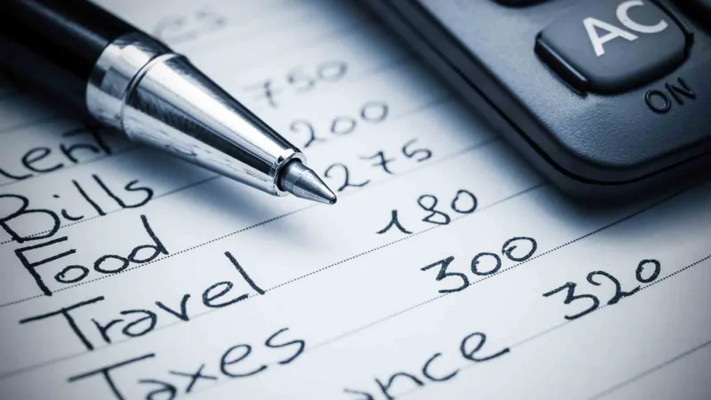 Pen resting on a handwritten household budget listing rent, bills, food, travel and taxes beside a calculator