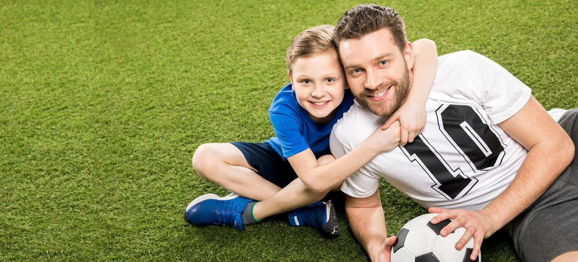 Father and son sitting on grass with a soccer ball, both smiling