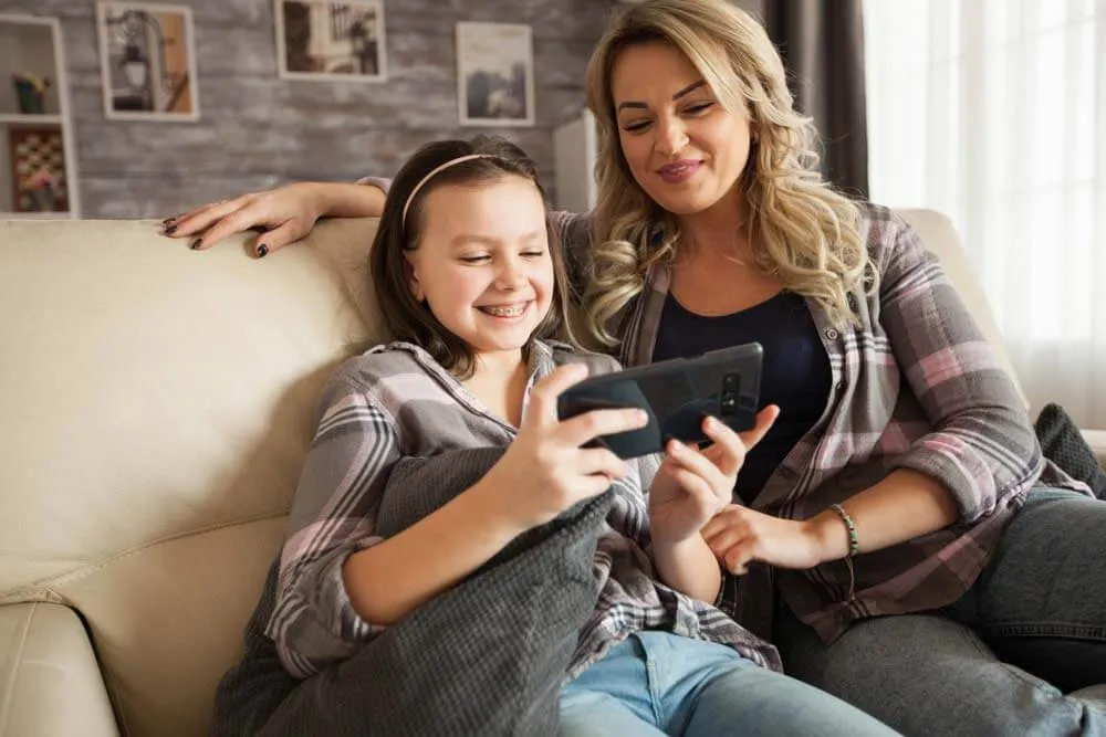 Mother and daughter sitting on a sofa laughing at a phone, the daughter wearing braces