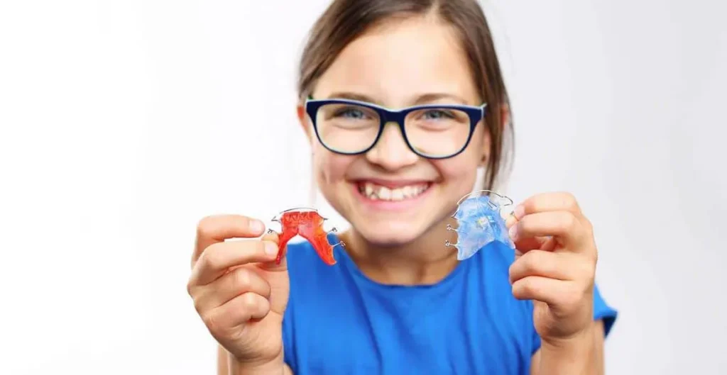 Young girl in glasses smiling and holding up two coloured orthodontic retainers