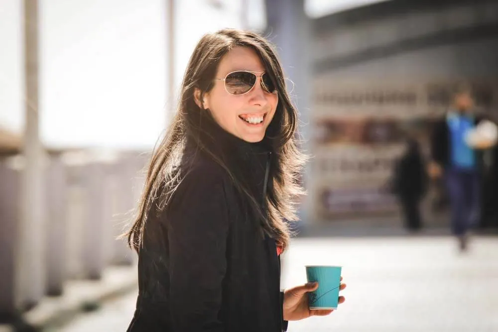 Woman in sunglasses smiling outdoors while holding a takeaway cup